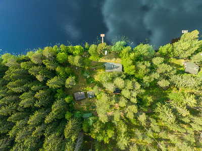 Kesämökkitontti ylhäältäpäin, taivas heijastuu lammen pinnasta ja puiden keskellä näkyy mökki ja piharakennukset. Kuvattu Urjalassa.